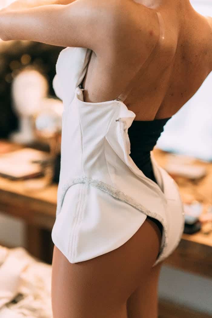 Back view of crop anonymous queer male putting on white bodysuit while preparing for performance in dressing room with table
